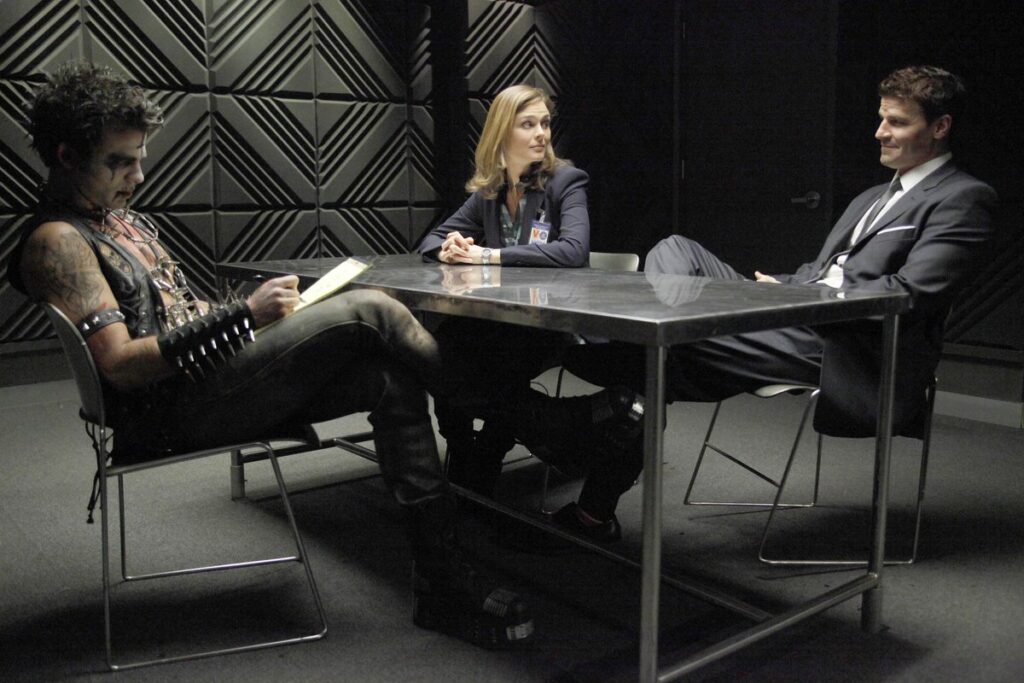 An interrogation room from the TV show Bones. Dr. Brennan and Agent Booth interrogate a man in a punk outfit. The lighting is warm and diffuse.