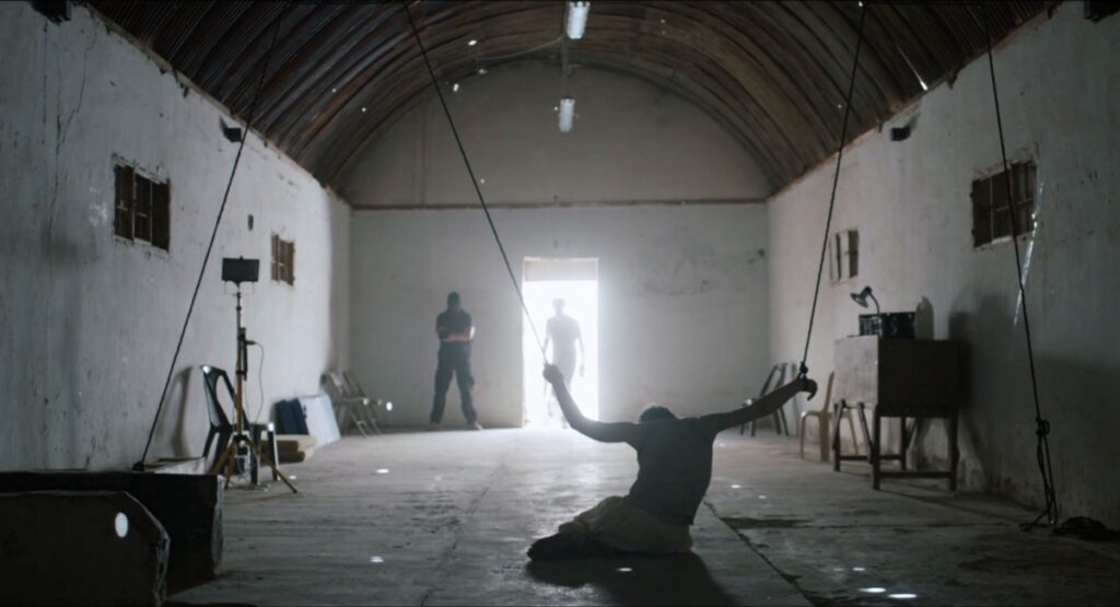 An interrogation about to begin from the movie Zero Dark Thirty. A prisoner kneels on the ground with his arms suspended by ropes from the cieling. In the middle of the shot, an interviewer enters from an overwhelmingly bright door.